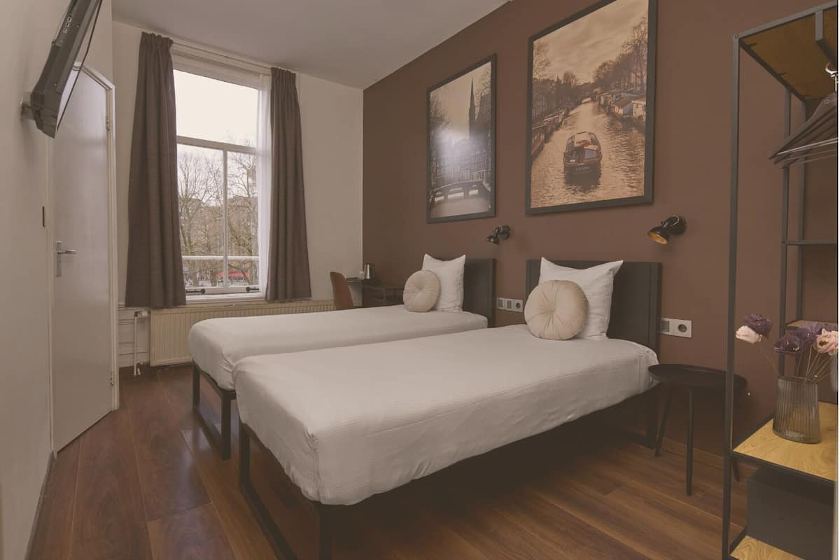A hotel room with two beds, a window with curtains, a radiator, a desk with a lamp, and two framed pictures on the wall.