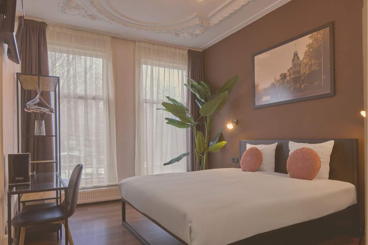 A hotel room with a large bed, a desk with a chair, a plant, and a painting on the wall.