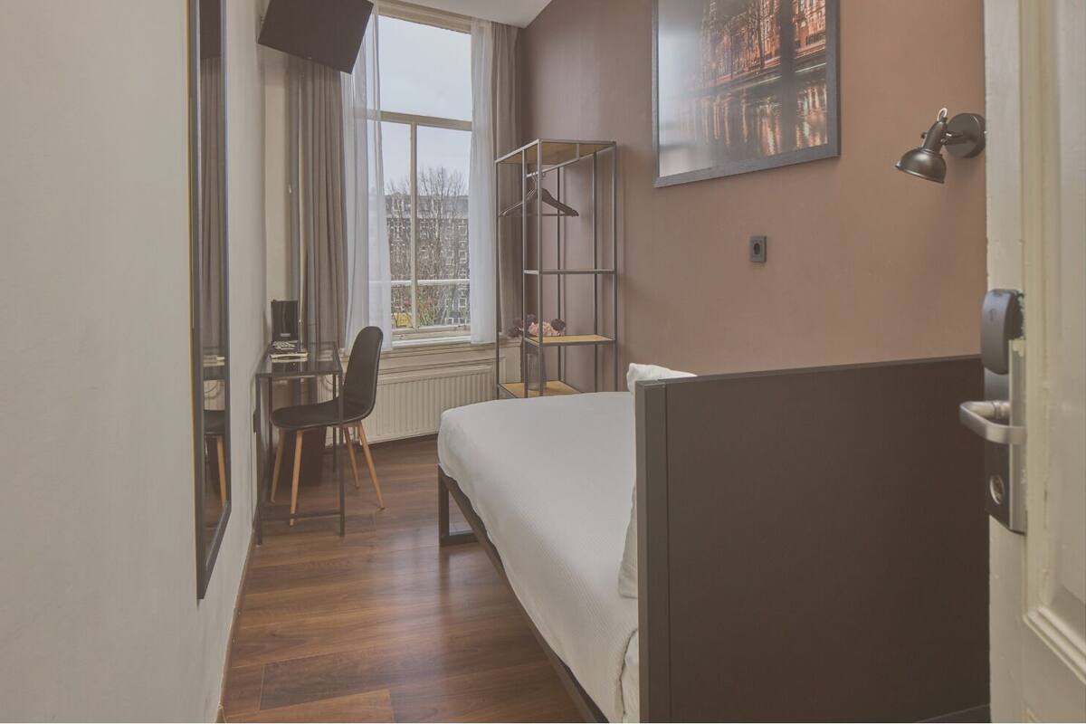 A hotel room with a bed, a desk with a chair, a window with a view of buildings, and a shelf with items.