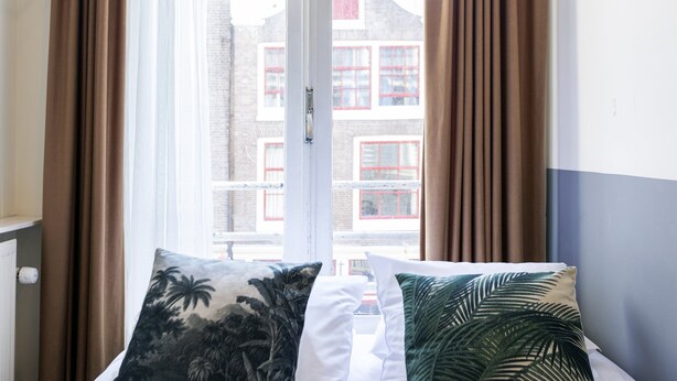Ein Bett mit zwei Kissen, ein Fenster mit Vorhängen und Blick auf Gebäude.