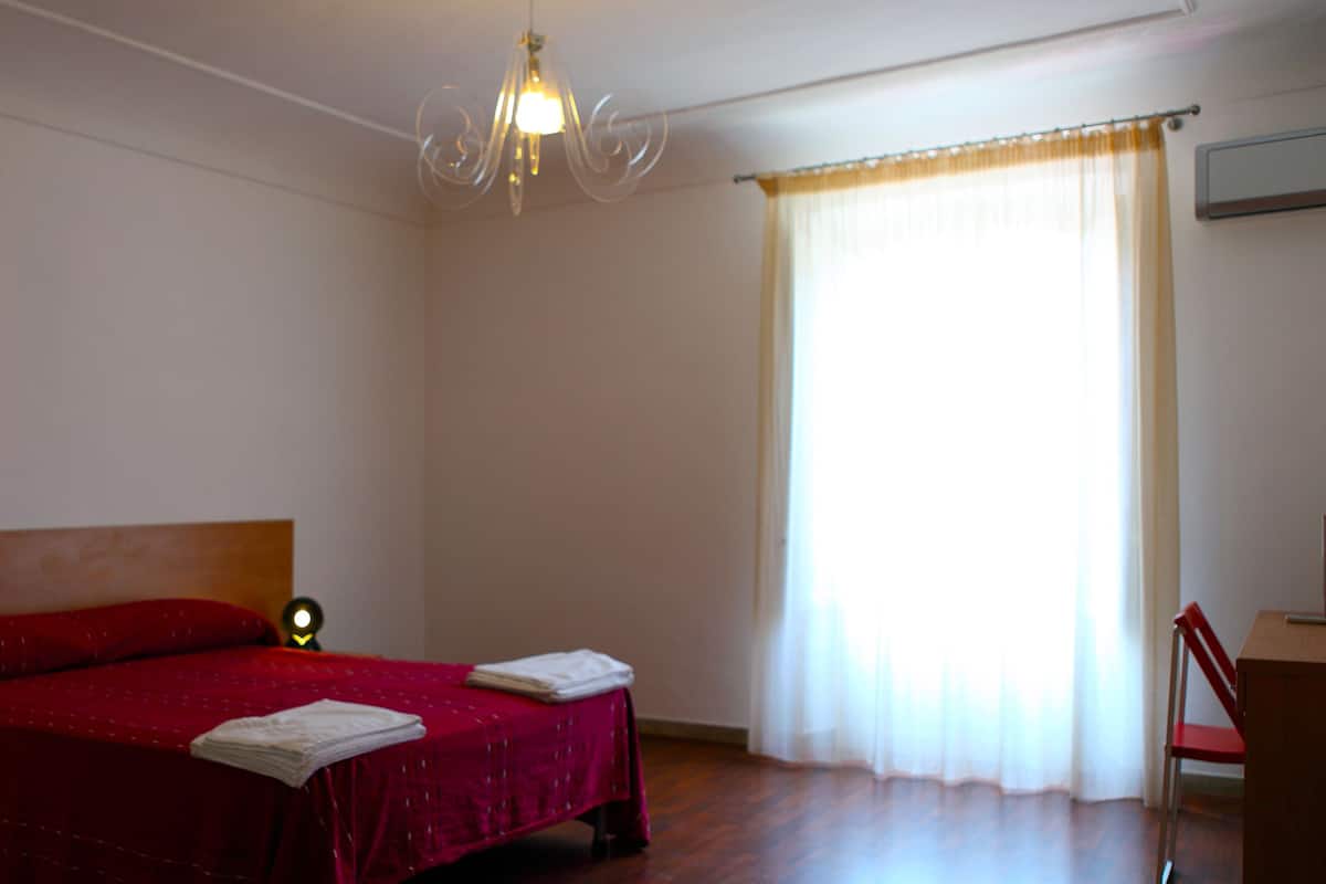 Une chambre avec une couvre-lit rouge, une tête de lit en bois, une lampe de chevet, une fenêtre avec des rideaux légers et un plancher en bois.