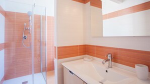 Un baño moderno con cabina de ducha de vidrio, un lavabo blanco con espejo y paredes con azulejos blancos y naranjas.