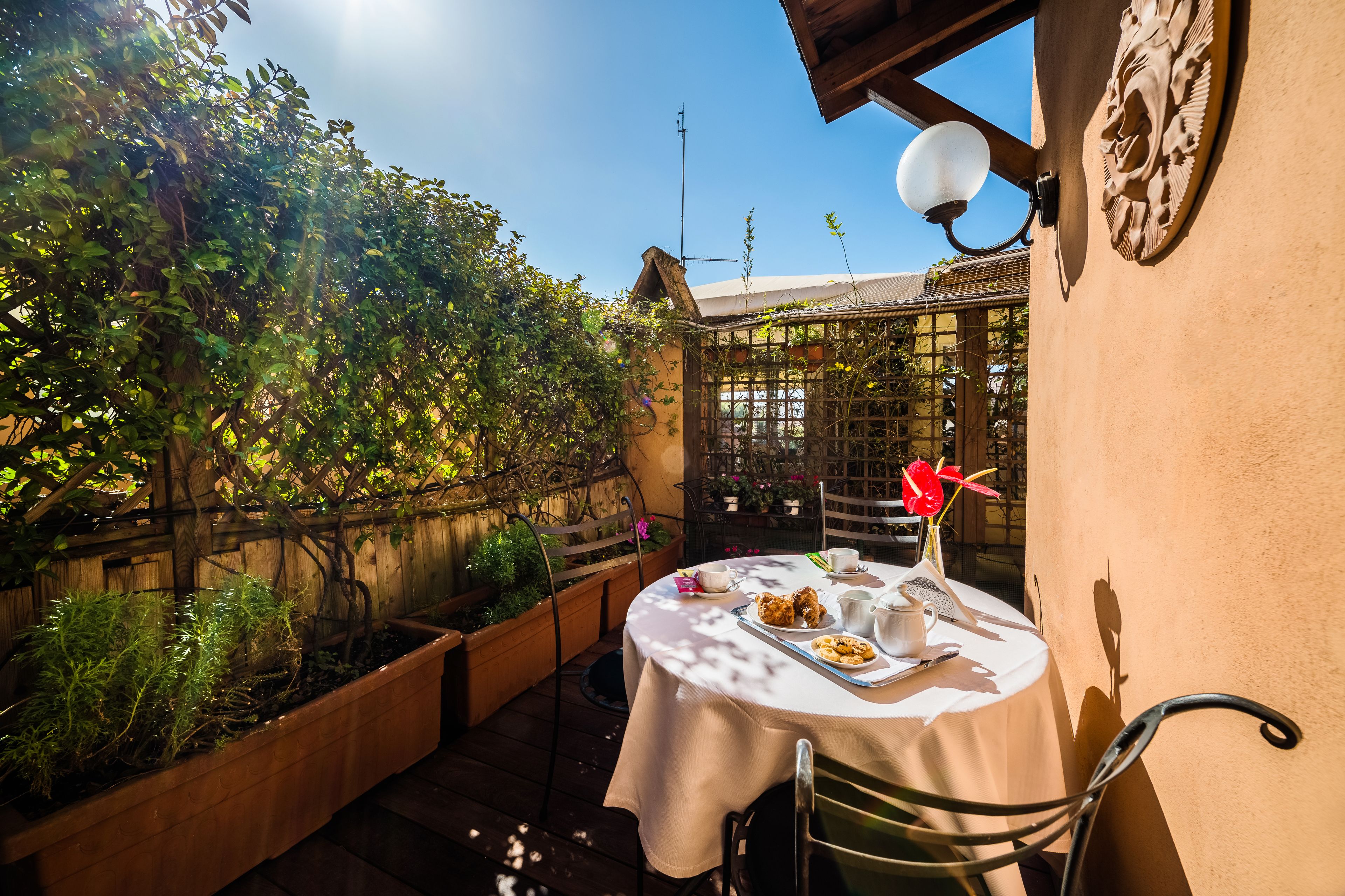 A round table with a white tablecloth, surrounded by chairs, set in a garden with wooden planters and a wooden fence.