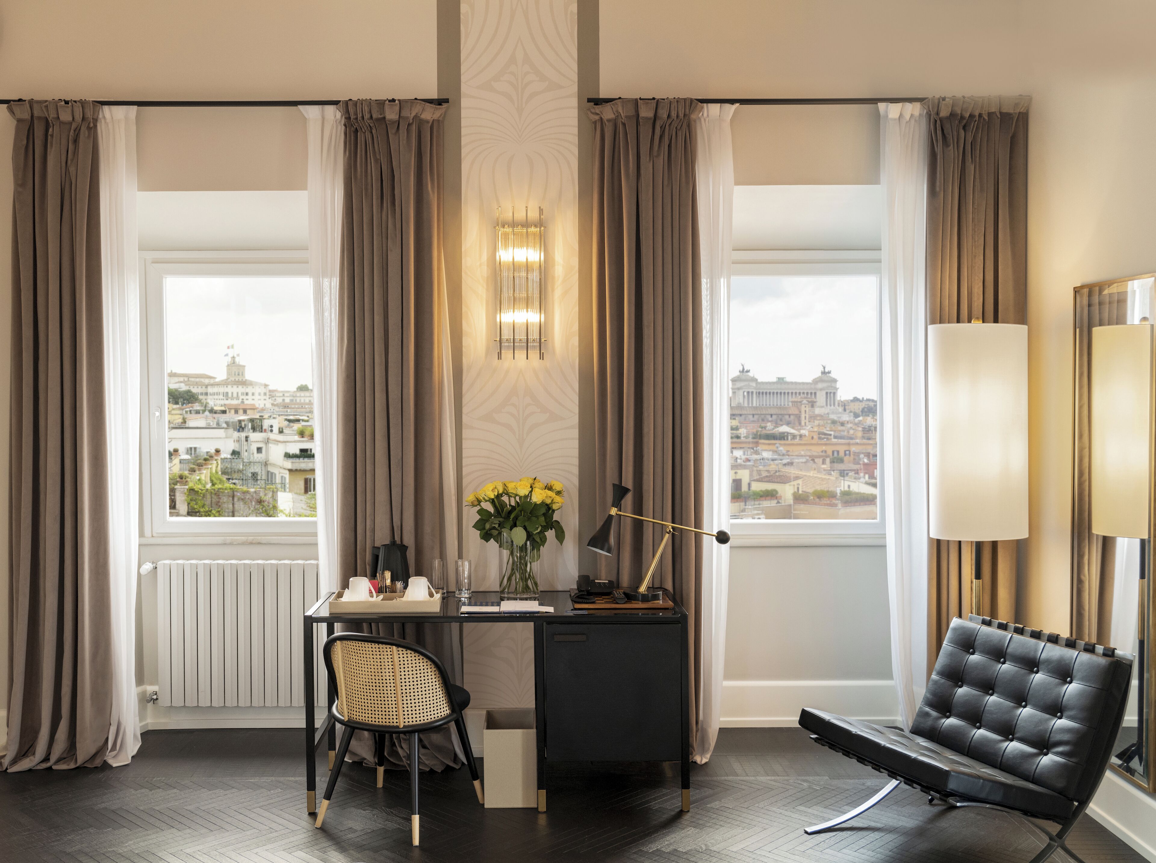 Habitación de hotel con escritorio, silla y sofá. Hay cortinas, una lámpara y vistas a la ciudad.