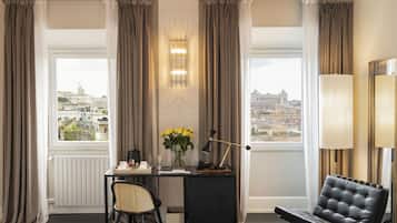 Habitación de hotel con escritorio, silla y sofá. Hay cortinas, una lámpara y vistas a la ciudad.
