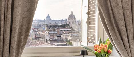 A room with a view of a cityscape, a dining table with food and drinks, and a vase of flowers.