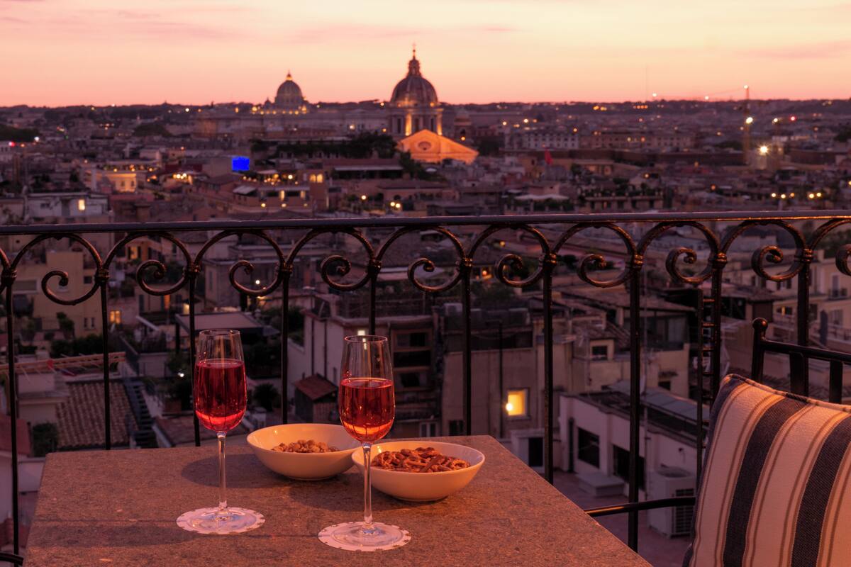 A balcony with two wine glasses and a bowl of nuts, overlooking a cityscape with domed buildings and a sunset sky.