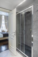 Una camera d'albergo moderna con doccia a parete in vetro, un letto con testiera scura e vista sulla città attraverso ampie finestre.