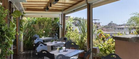 An outdoor patio with wooden tables and chairs, surrounded by potted plants and a view of the cityscape.