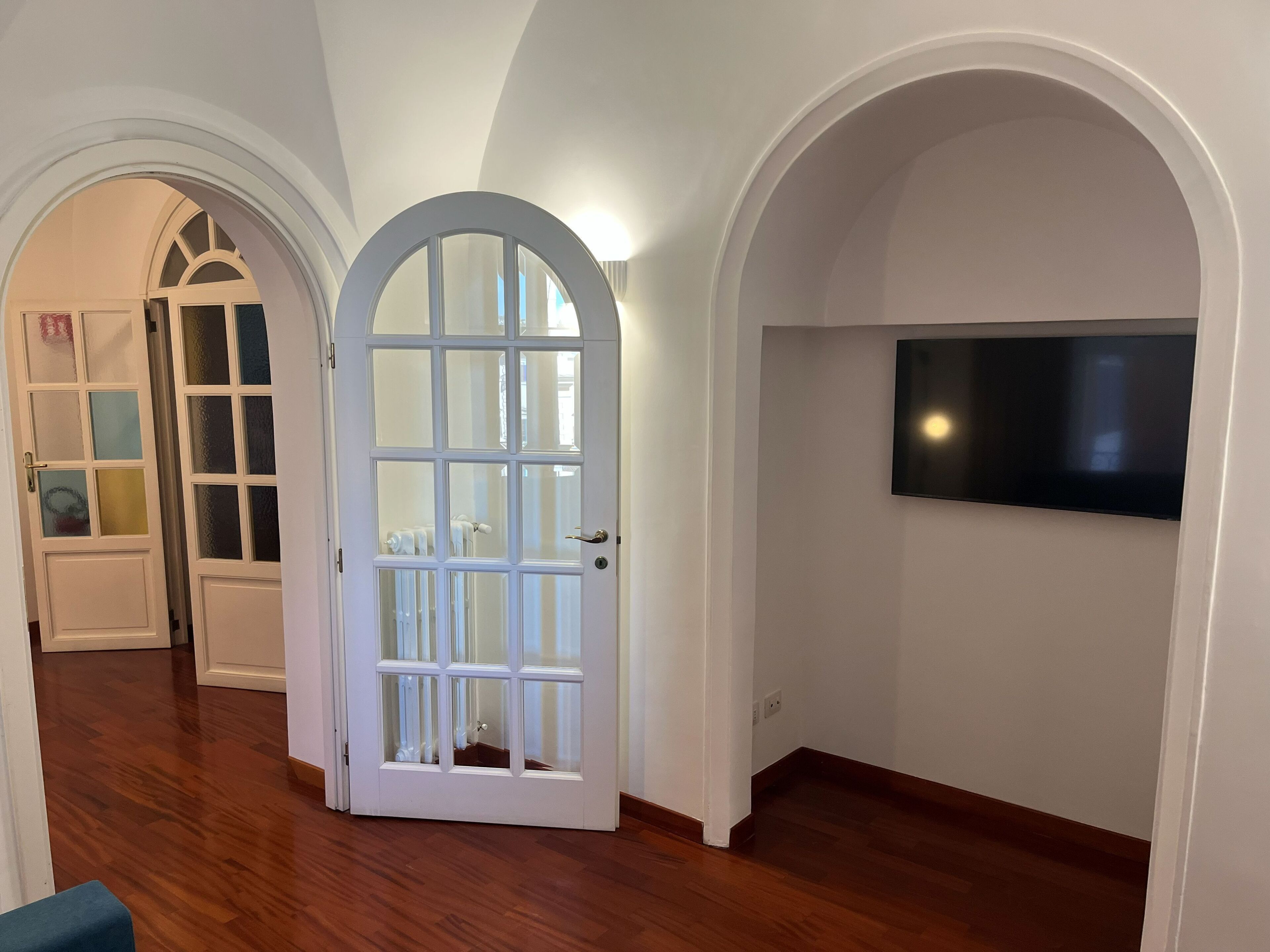 Un pasillo con una puerta blanca, piso de madera y un televisor montado en la pared.