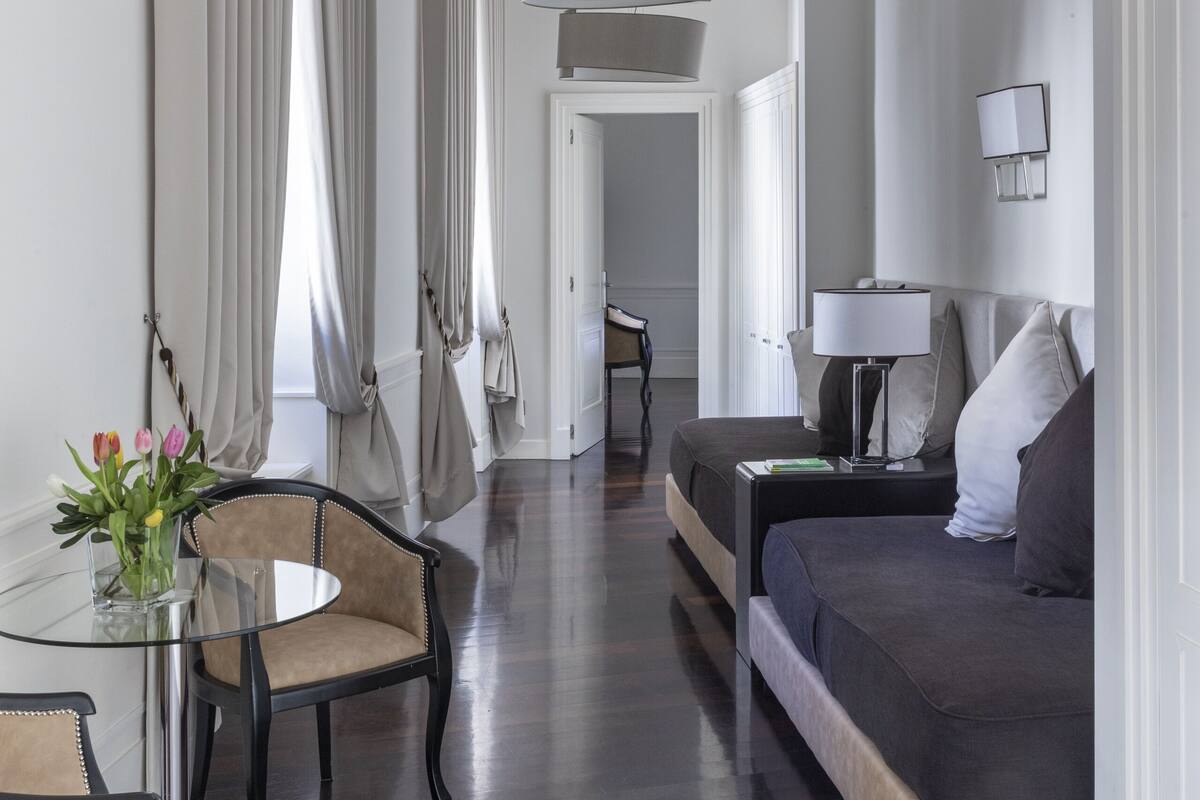 A modern hotel room with a dark wood floor, a glass table with chairs, a sofa, and a large window with curtains.
