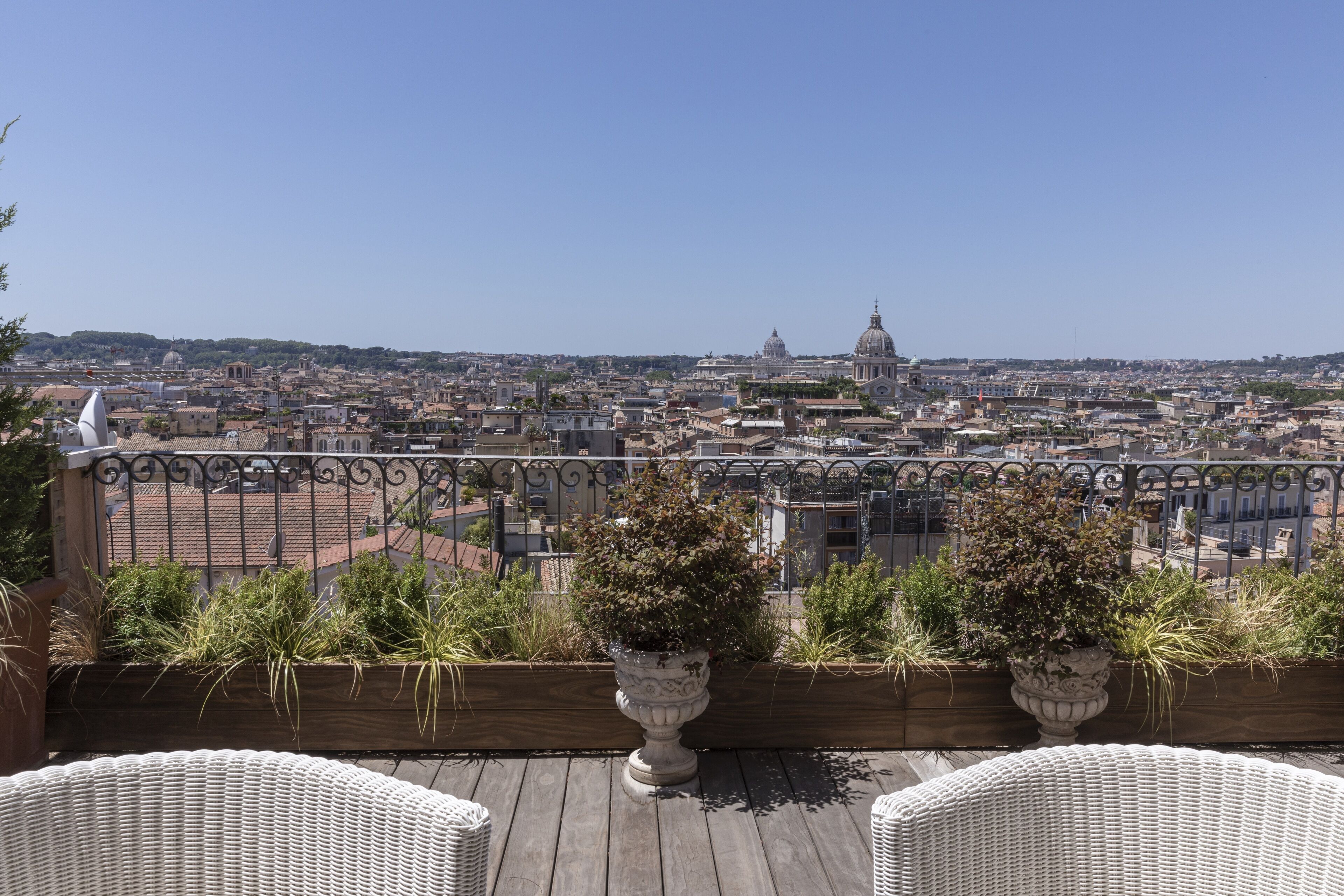 En balkon med fletstole og krukker med planter, med udsigt over en by med historisk arkitektur og en klar himmel.