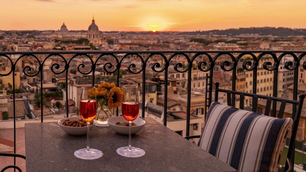 A balcony with a table set for two, featuring wine glasses and a bowl of snacks, overlooking a cityscape with a prominent dome building in the distance during sunset. - Hotel Scalinata di Spagna (Rome)
