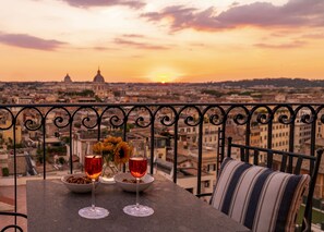 A balcony with a table set for two, featuring wine glasses and a bowl of snacks, overlooking a cityscape with a prominent dome building in the distance during sunset. - Hotel Scalinata di Spagna (Rome)