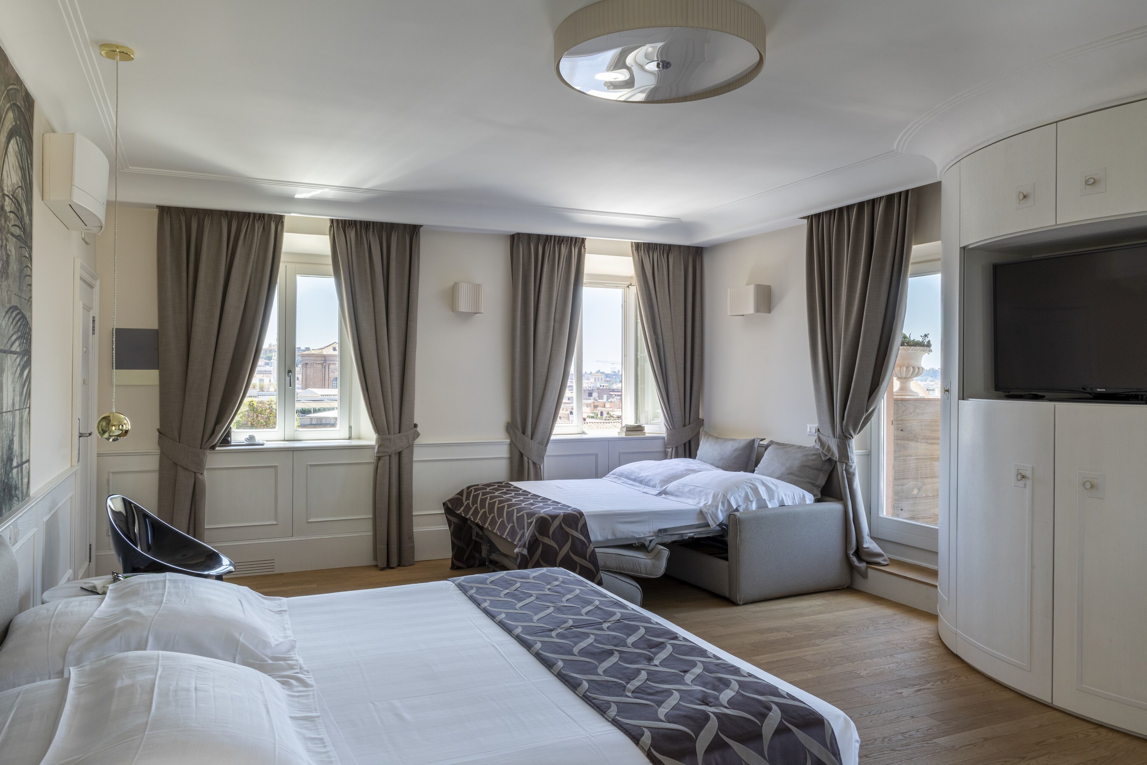 A hotel room with a bed, a sofa bed, a TV, and a view of the city.