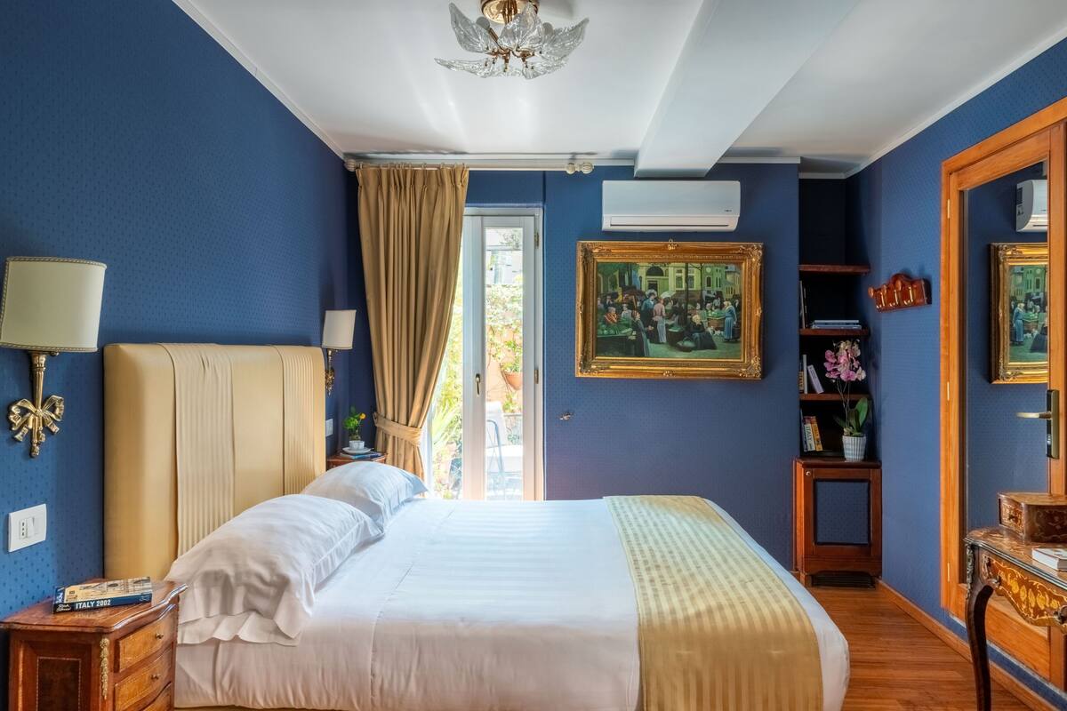 A bedroom with a large bed, a nightstand, a dresser, a ceiling fan, a window with curtains, and a framed painting on the wall.