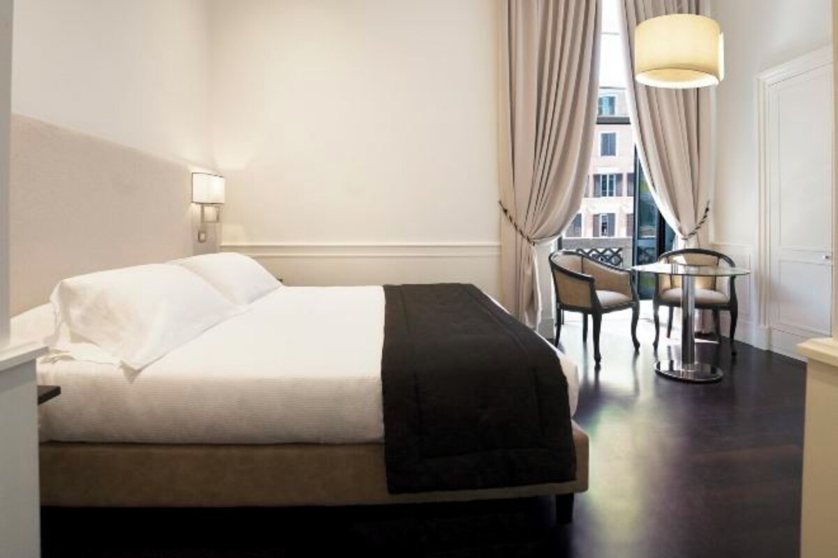 A hotel room with a bed, a desk with chairs, a lamp, and a view through curtains.