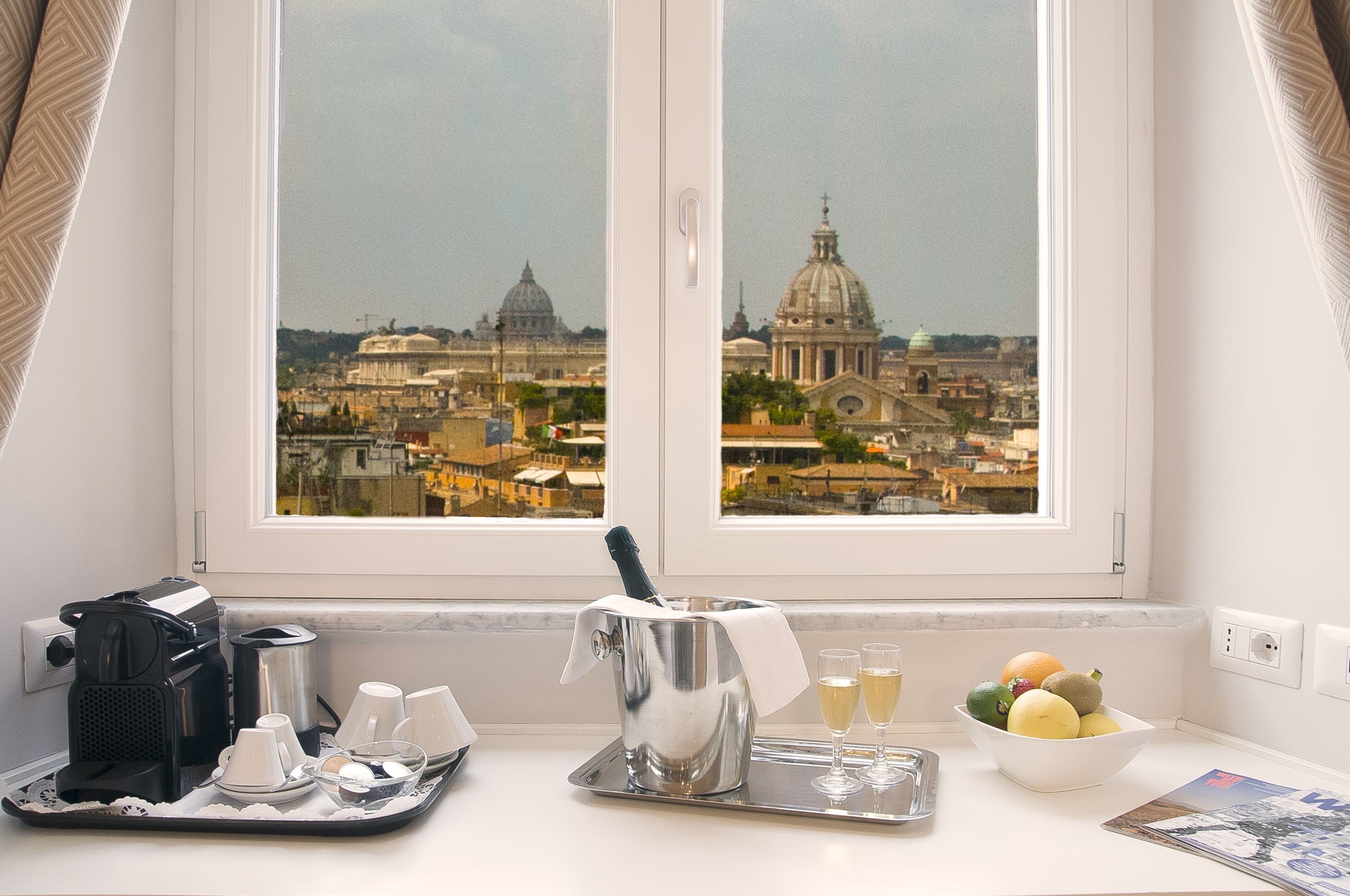 A kitchen with a view of a cityscape, a coffee machine, a silver pot, glasses, and a bowl of fruit.