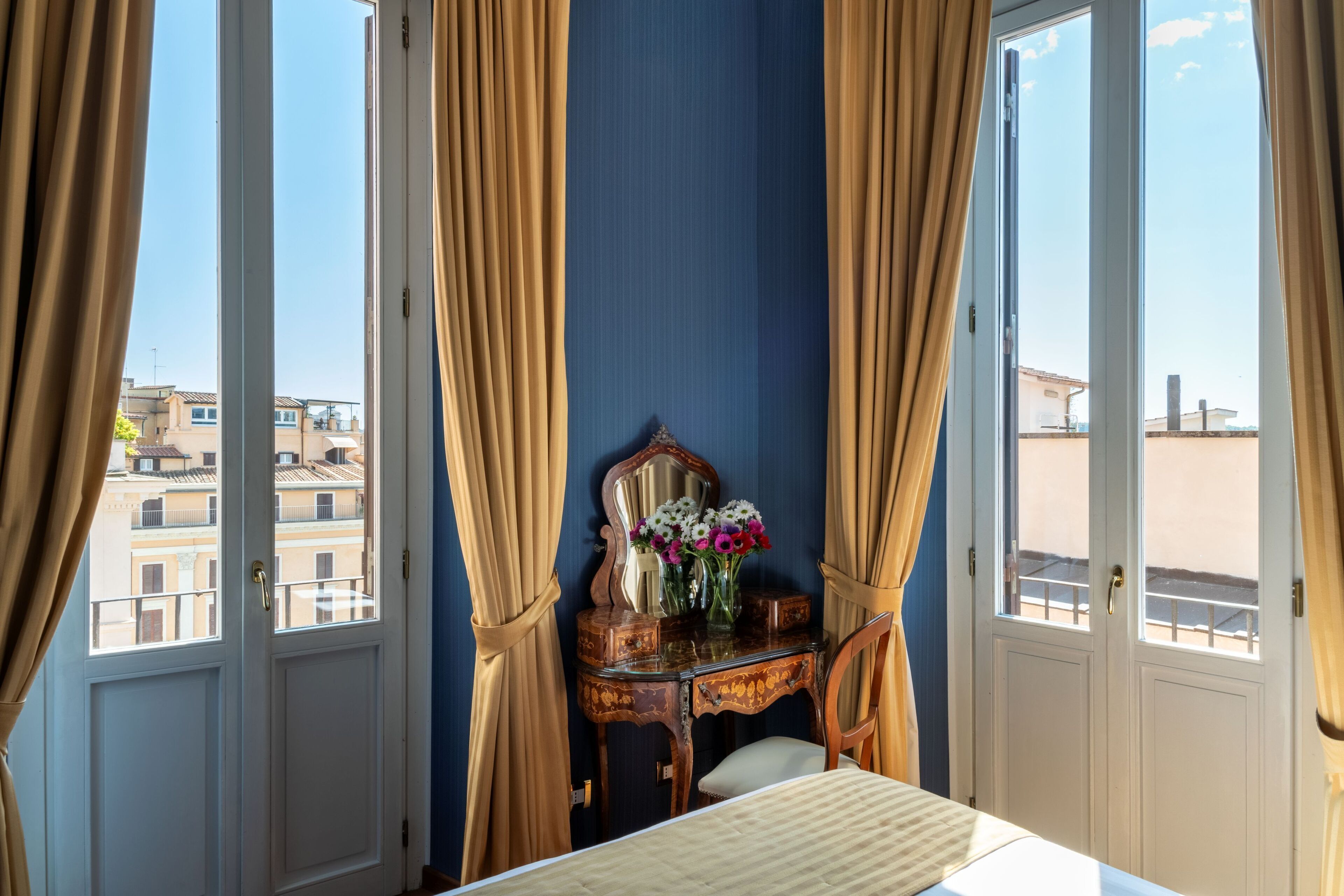 A hotel room with a bed, a vanity with a mirror, and a vase of flowers. There are curtains on the windows, and a view of buildings outside.