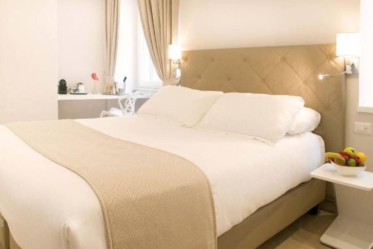 A hotel room with a large bed, a bedside table with a fruit bowl, and a desk with a lamp.