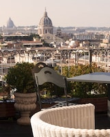 Un balcone con una sedia in vimini, una sedia in metallo e una pianta in vaso. Sullo sfondo, si vede un paesaggio urbano con edifici a cupola.