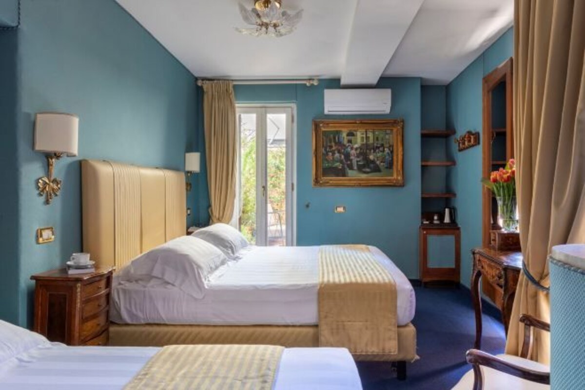 A hotel room with two beds, a window with curtains, a painting on the wall, and a desk with a chair.