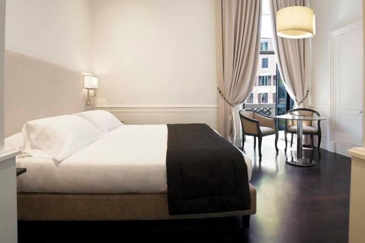 A hotel room with a bed, a desk with chairs, a lamp, and a view through curtains.