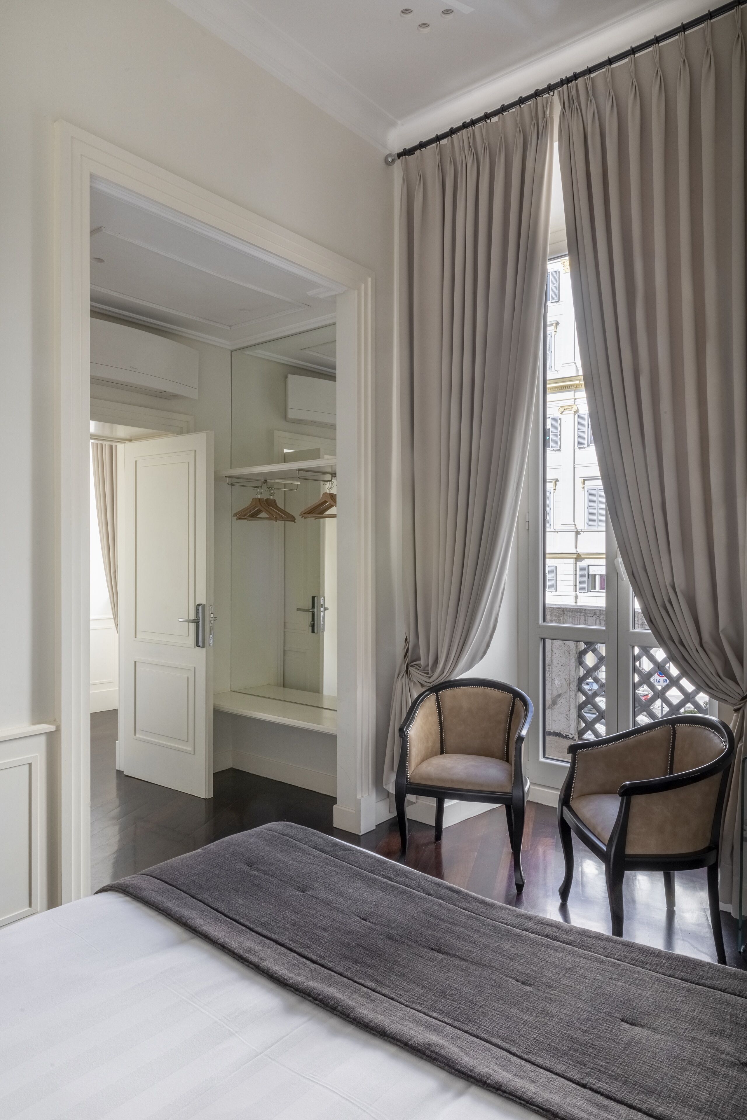 A hotel room with a bed, two armchairs, a large mirror, and a view of the street through a window with curtains.