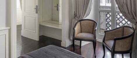 A hotel room with a bed, two armchairs, a large mirror, and a view of the street through a window with curtains.