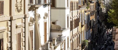 A narrow street lined with multi-story buildings, featuring balconies and a row of parked cars.