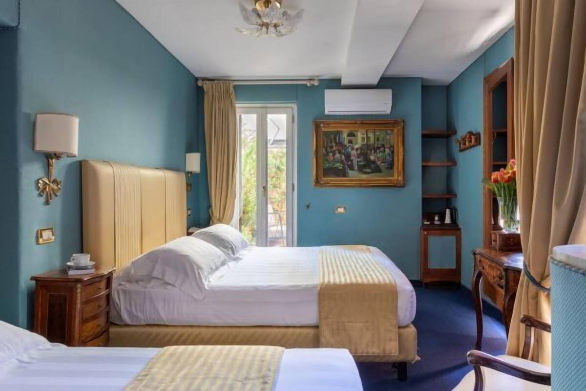 A hotel room with two beds, a window with curtains, a painting on the wall, and a desk with a chair.