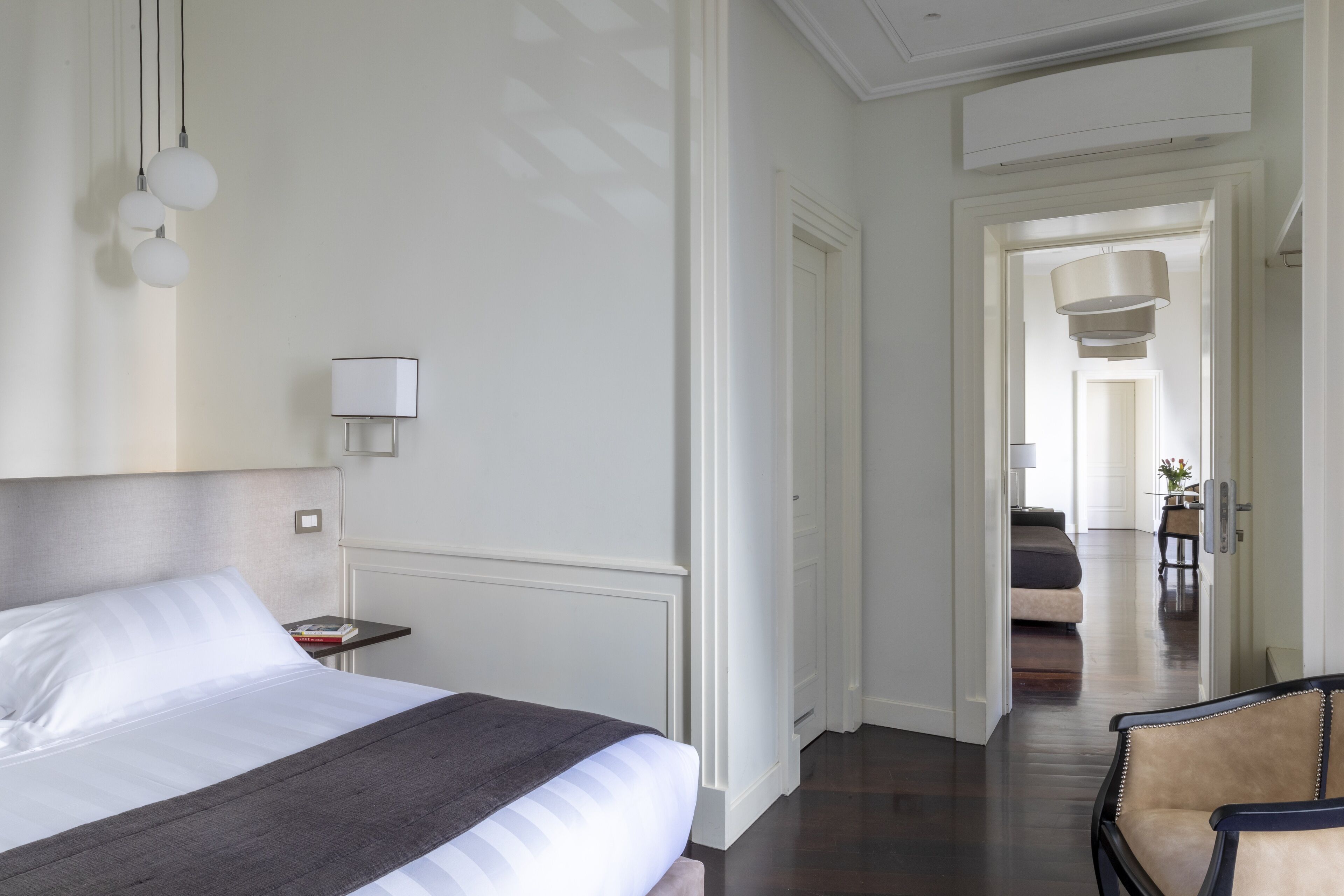 Ein Hotelzimmer mit einem großen Bett, Nachttischen, einem Sessel und einem Gang zu einem weiteren Zimmer.