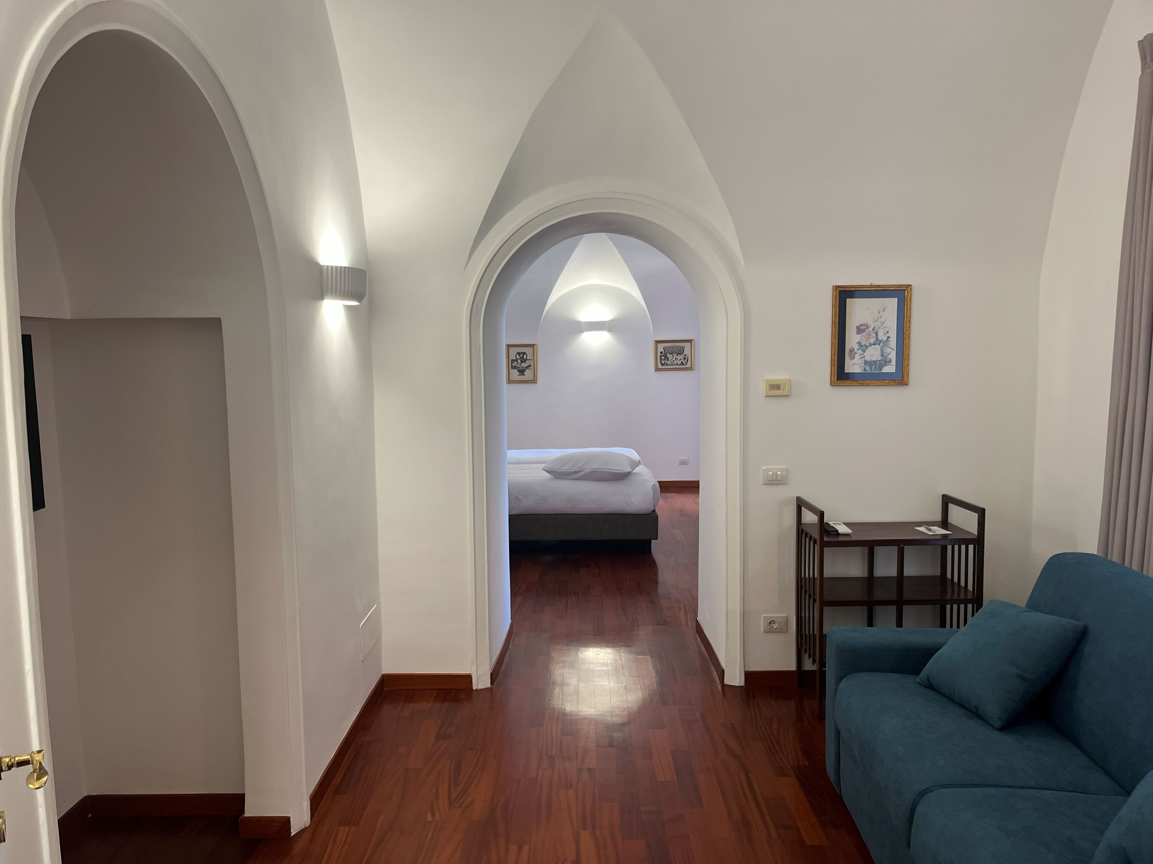 A hallway leading to a bedroom with a bed, a blue sofa, and framed artwork on the walls.