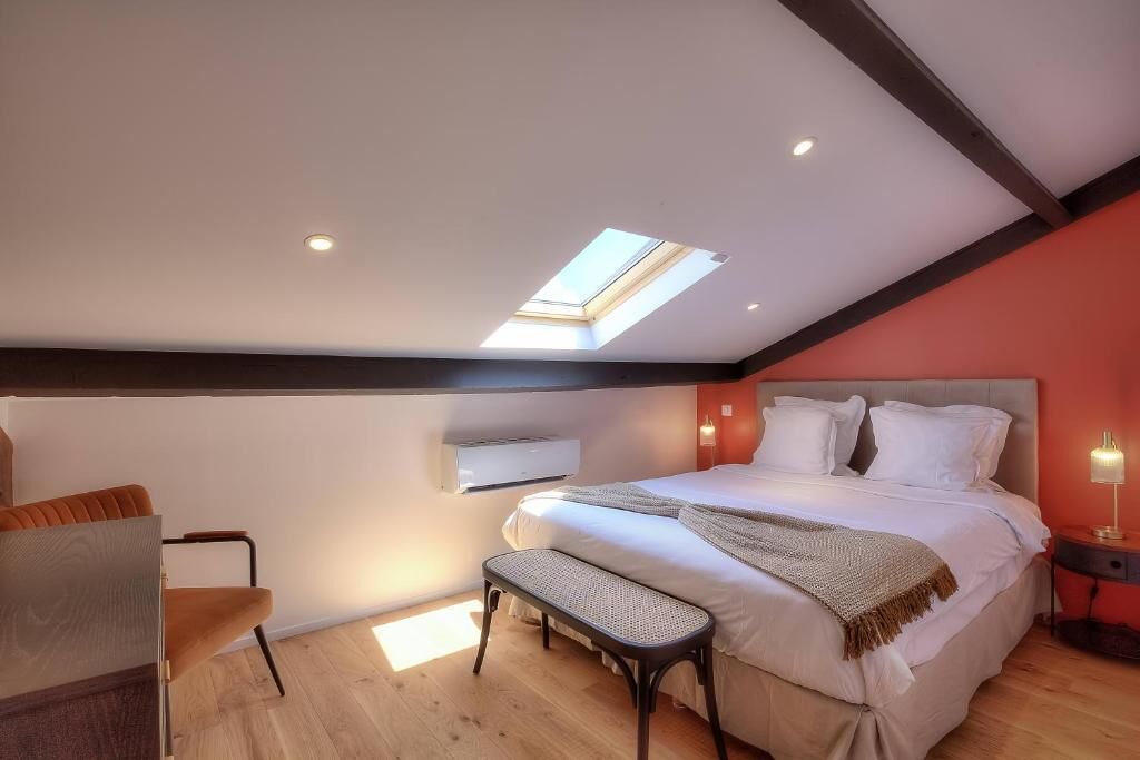 A bedroom with a skylight, a bed with white linens, a bench, and a bedside table with a lamp.