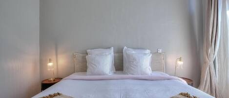 A neatly made bed with white bedding and two bedside lamps, a beige throw blanket, and a plain wall with a curtain on the right.