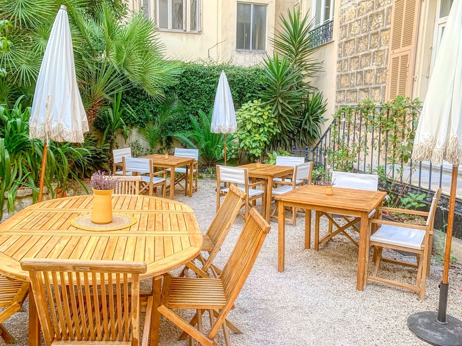 Un espace patio aménagé avec des tables et des chaises en bois, entouré d’une végétation luxuriante et d’un mur en pierre.