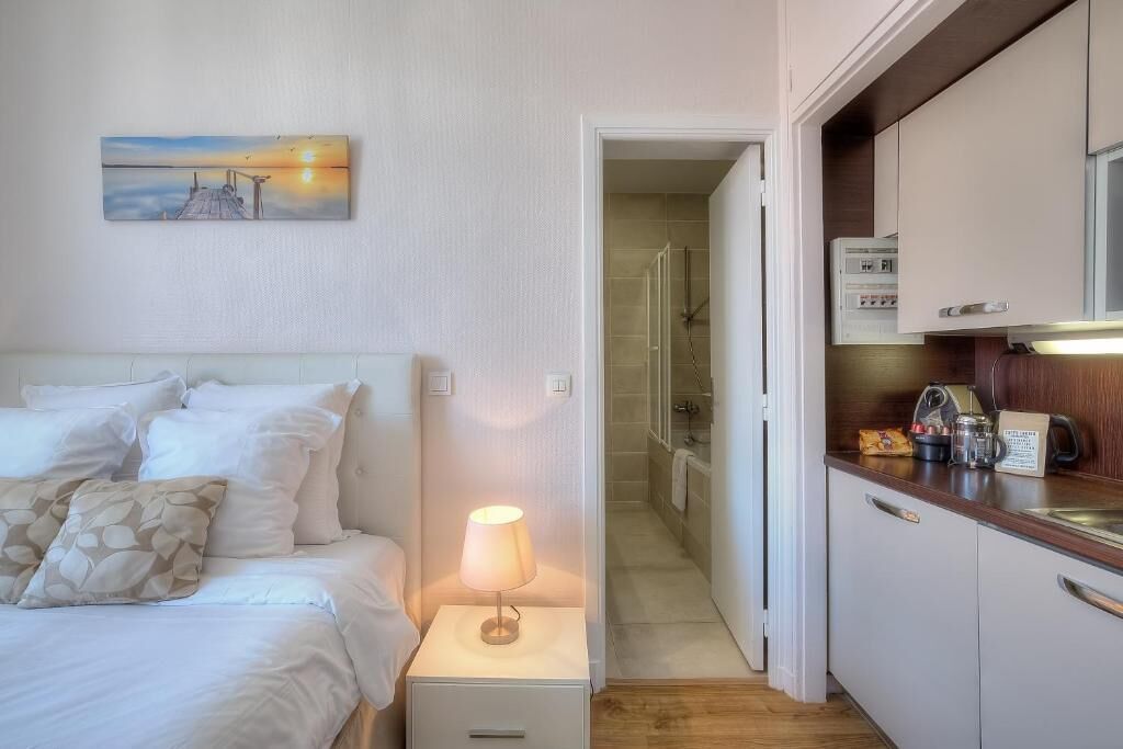 A hotel room with a bed, bedside lamp, a painting on the wall, a kitchenette, and a bathroom visible through an open door.