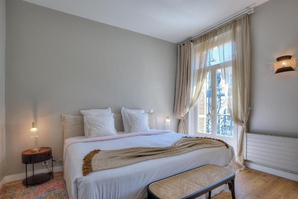 Un dormitorio con cama, lámparas de noche, una ventana con cortinas y un radiador.