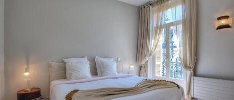 Un dormitorio con cama, lámparas de noche, una ventana con cortinas y un radiador.