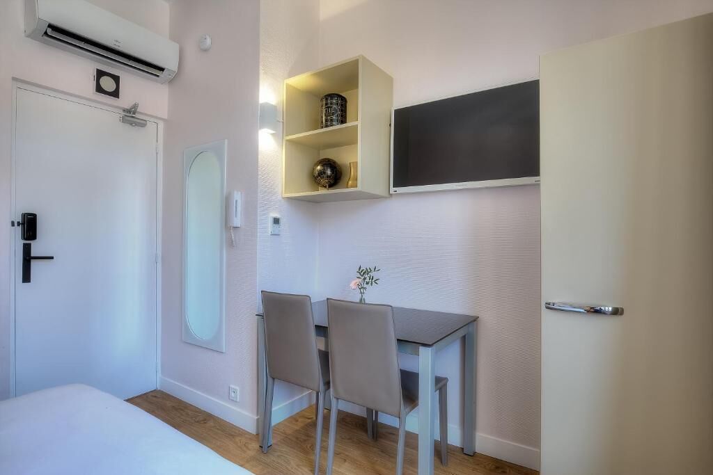 A compact room with a bed, a desk with two chairs, a small shelf with decorative items, a wall-mounted TV, and an air conditioning unit.