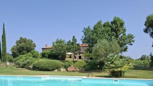 A swimming pool with a clear blue water feature, surrounded by a brick patio. In the background, there is a stone building with a tiled roof, surrounded by lush greenery and trees. - Tenuta di Seripa (Sassetta)