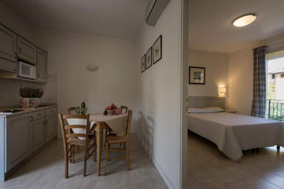 Una habitación compacta con kitchenette, un comedor con mesa y sillas, y una cama individual con mesita de noche.