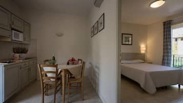 A compact room with a kitchenette, a dining area with a table and chairs, and a single bed with a nightstand.