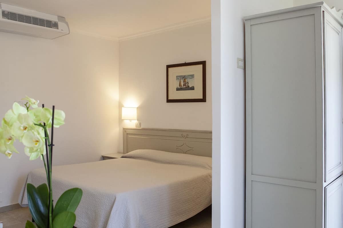 Una habitación de hotel con cama, mesita de noche, lámpara, un cuadro en la pared y un armario blanco.