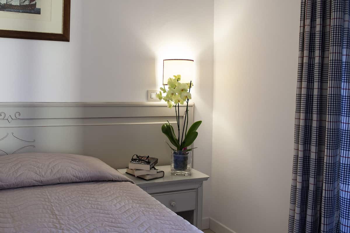 Una cama bien hecha con cubrecama acolchado, una mesita de noche con lámpara, un cuadro enmarcado y una orquídea en maceta.