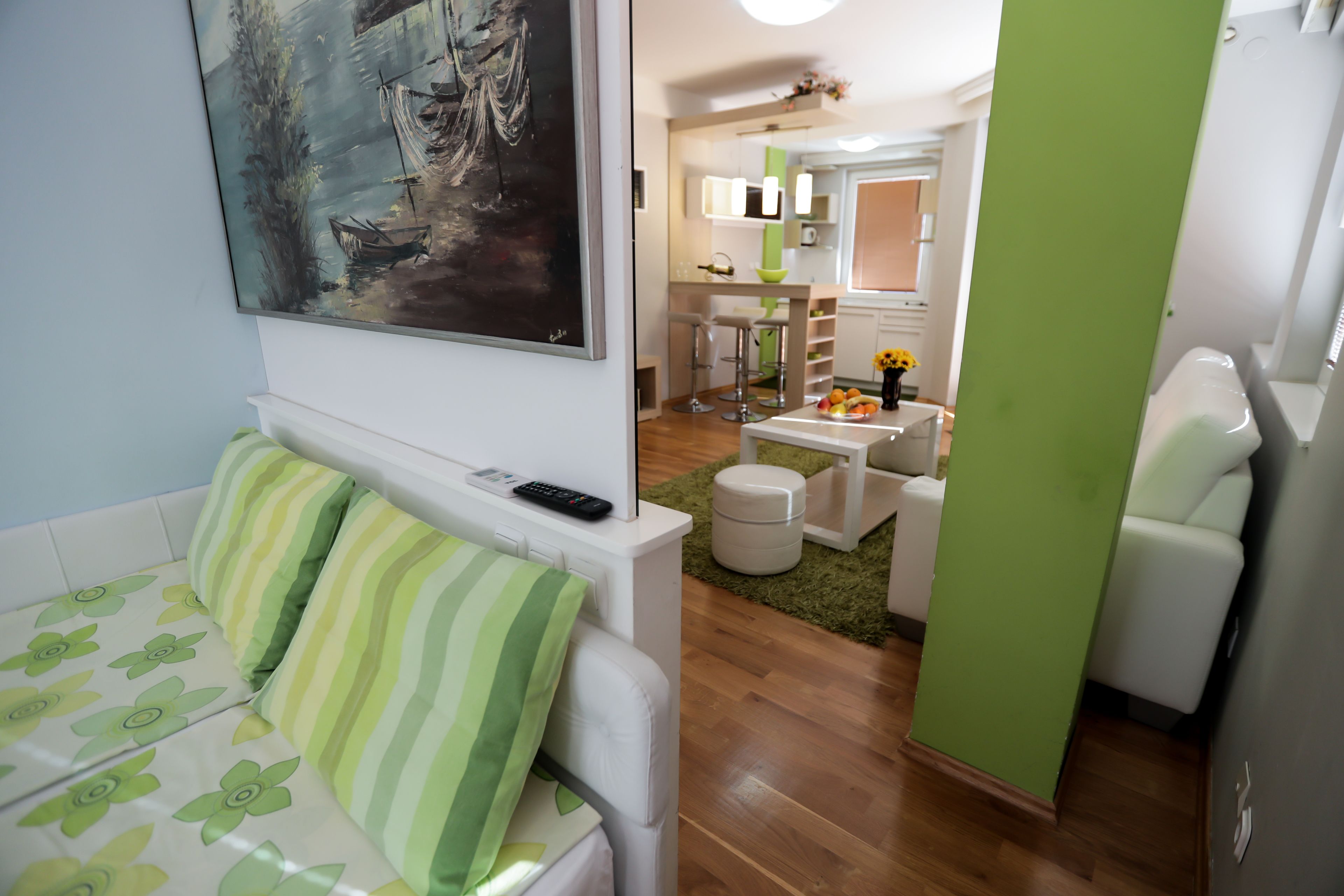 A modern living room with a sofa, a painting, a remote control, and a kitchen area in the background.