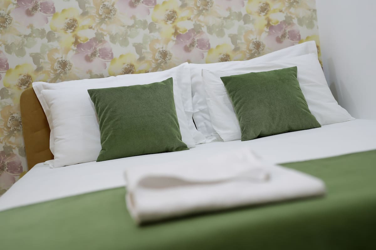 A neatly made bed with white and green pillows, a folded white towel, and a floral wallpaper background.
