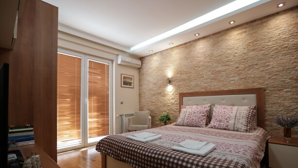 Un dormitorio con una colcha a cuadros, un cabecero de madera, una mesita de noche con un florero y un ventanal grande con persianas.