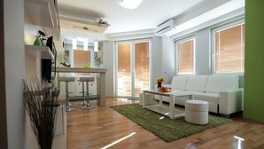 Una sala de estar moderna con un sofá blanco, una mesa de centro y un tapete verde. Se observa una zona de cocina con una barra y una mesa de comedor.