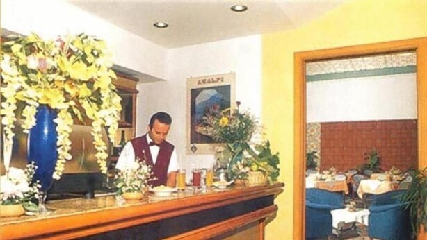 Zona de recepción de hotel con un mostrador de madera, una recepcionista y un área de comedor con mesas y sillas.